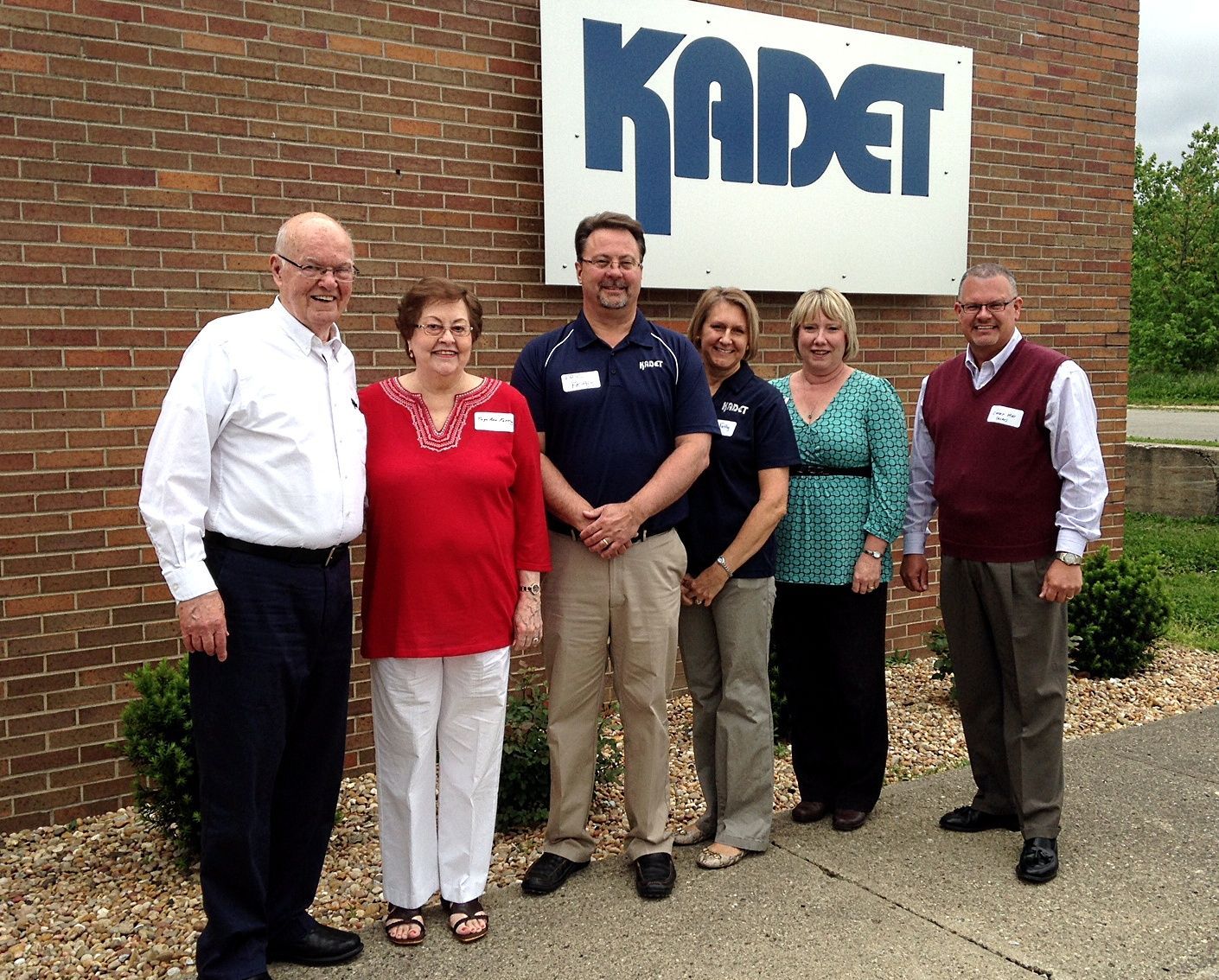 Six people stand in a line smiling in front of a brick building with a large sign that reads