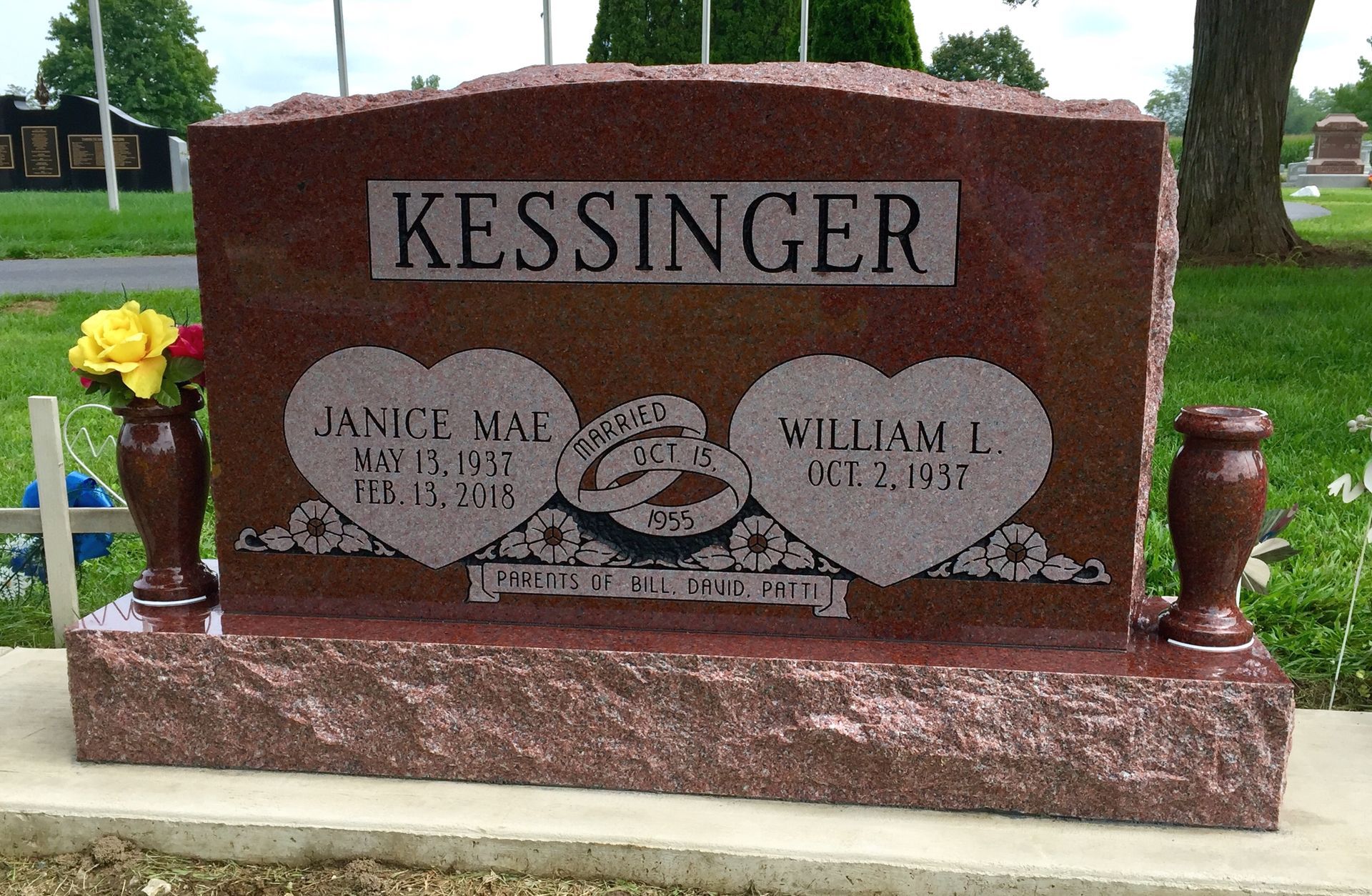 A red granite Kessinger double headstone featuring two hearts, a banner with rings, and side vases in a grassy cemetery.