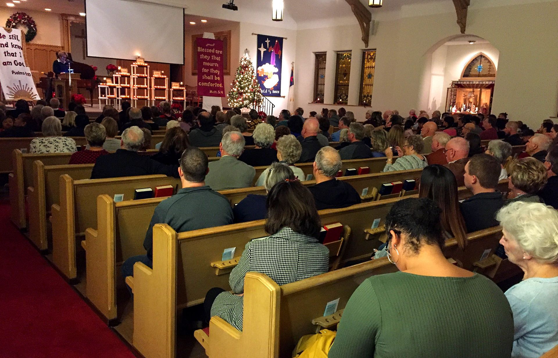 A congregation sits in wooden pews facing a stage in a church during a service, with a screen and decorations visible.