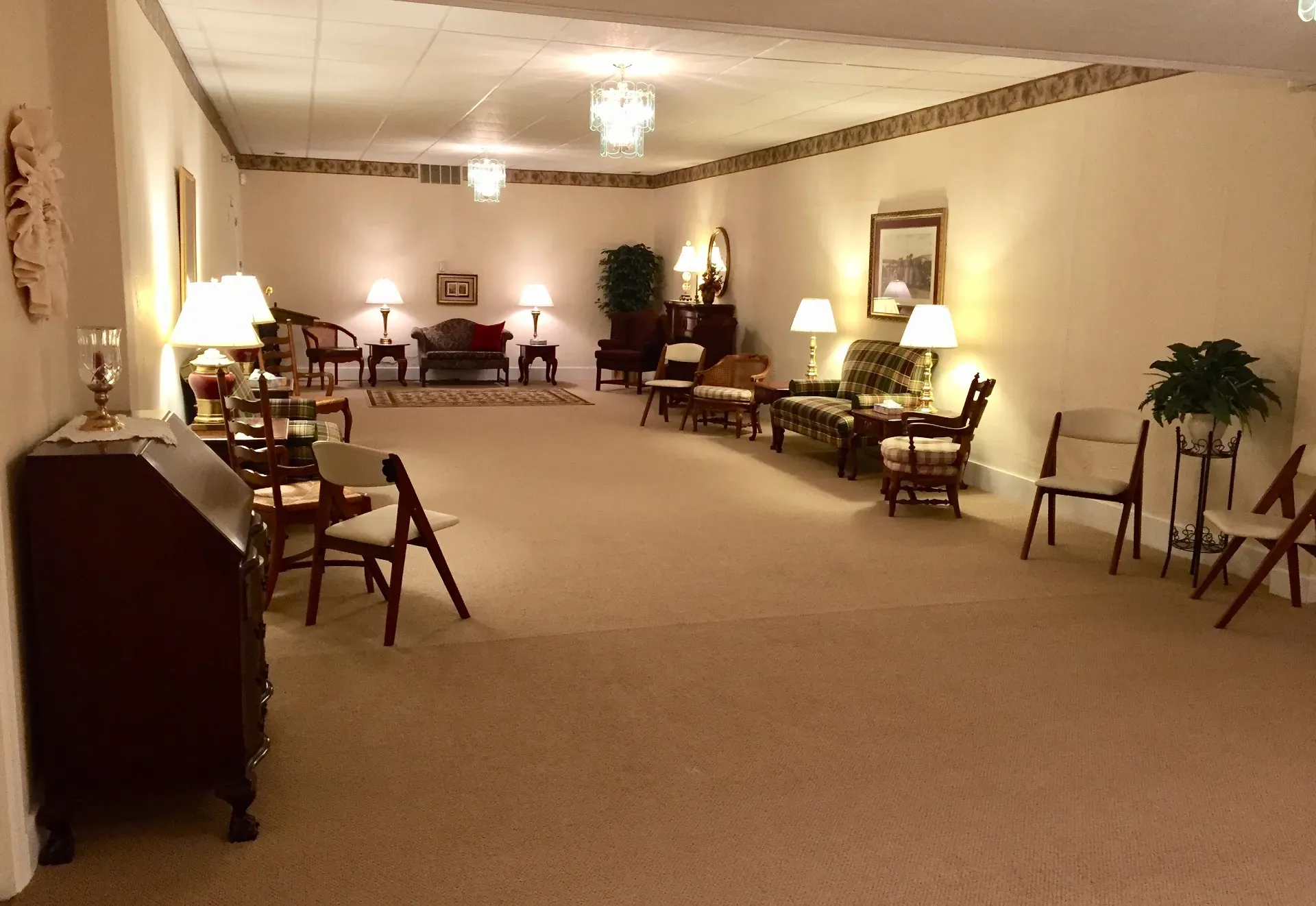 A wide view of a beige-carpeted funeral parlor waiting room furnished with chairs, side tables, and warm lamps.