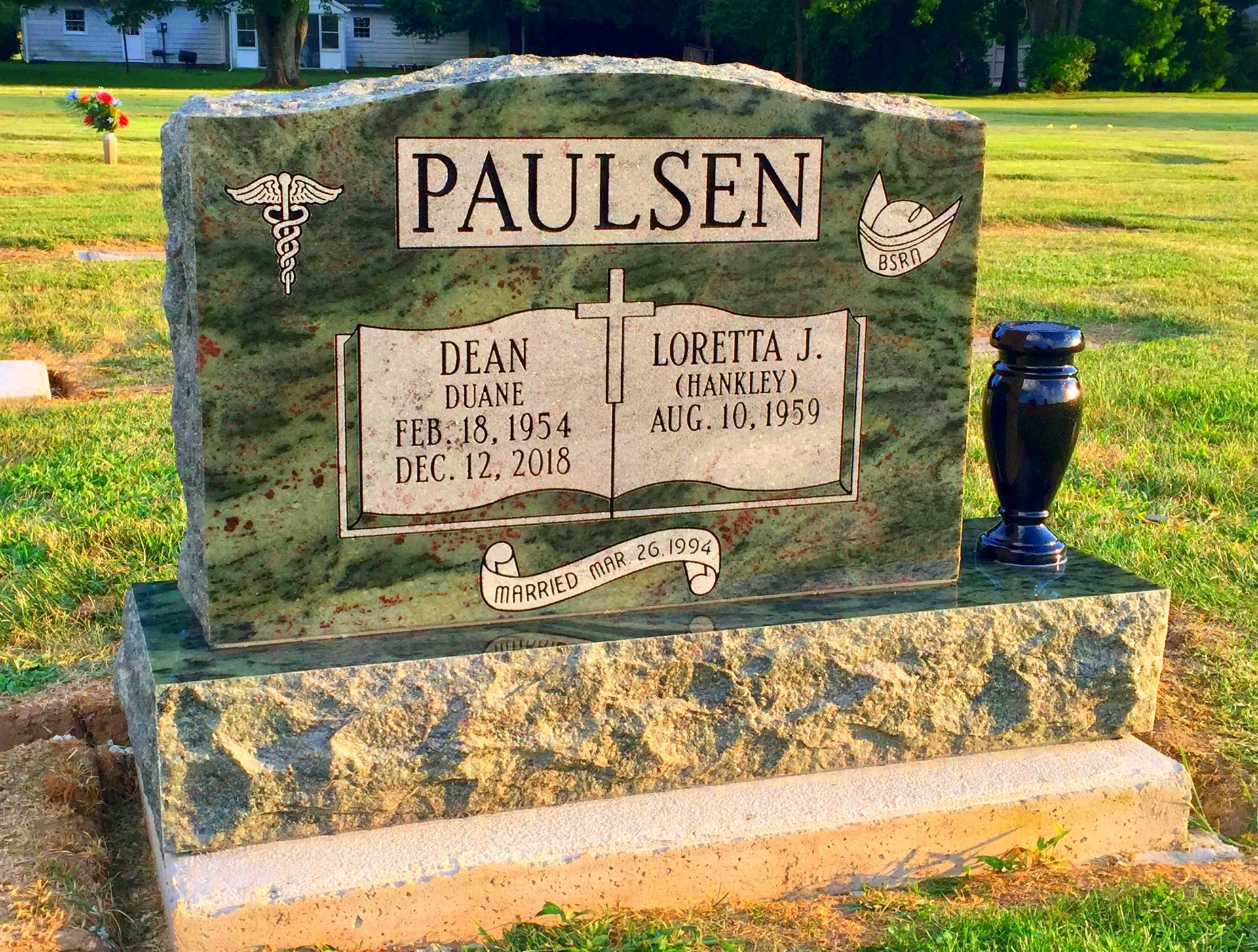 A green granite Paulsen family tombstone with an engraved open book, a cross, and a black vase in a grassy cemetery.