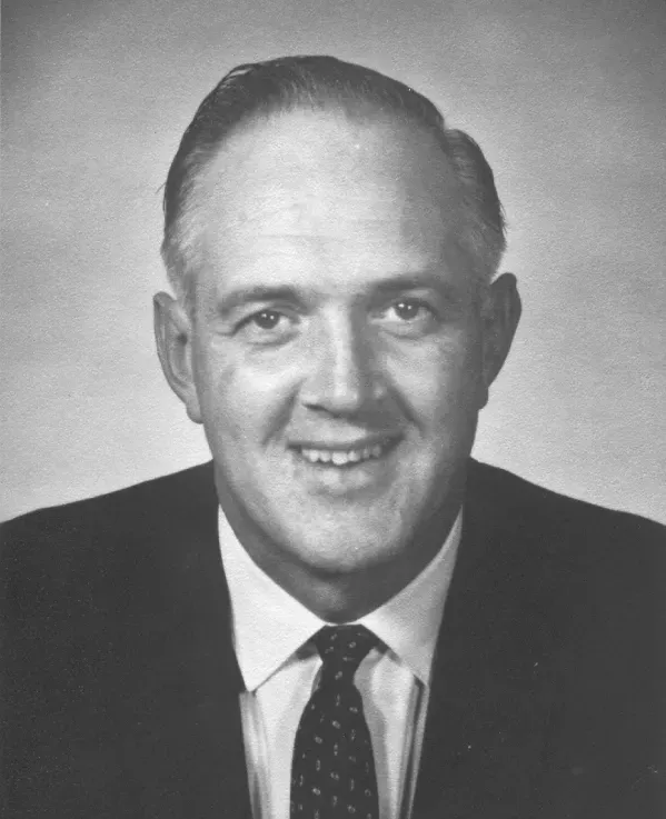 A studio head-and-shoulders portrait of a man in a dark suit and patterned necktie against a plain, light background.