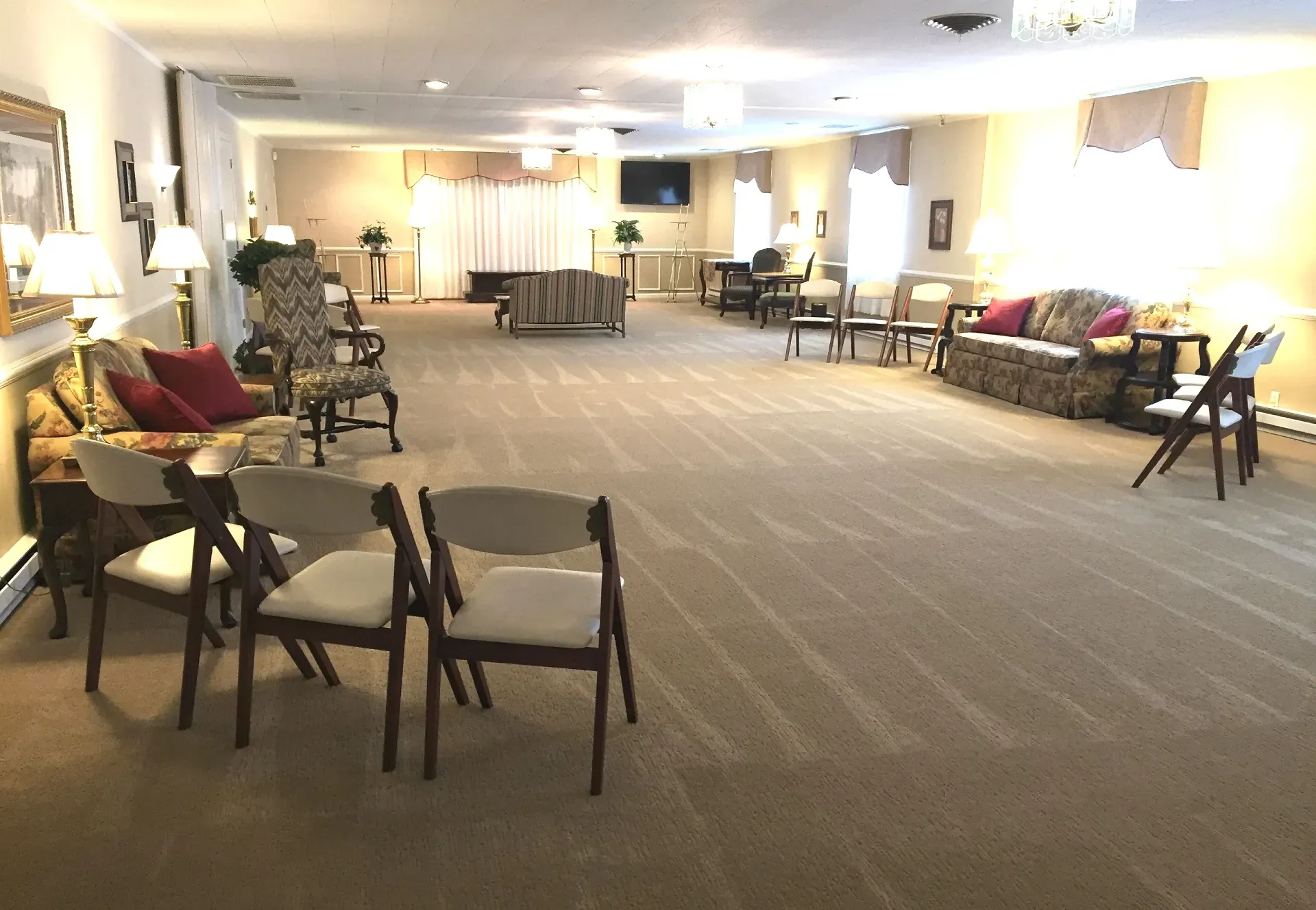 A wide, carpeted room set up for an event with chairs, sofas, and lamps, featuring a television in the far back.