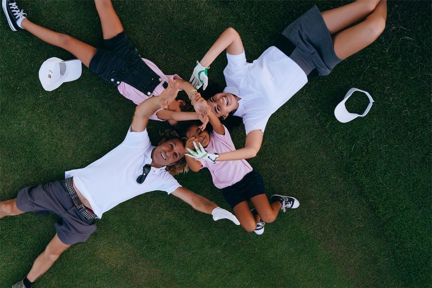 Six people lie in a circle on grass, stacking hands together in a team huddle.