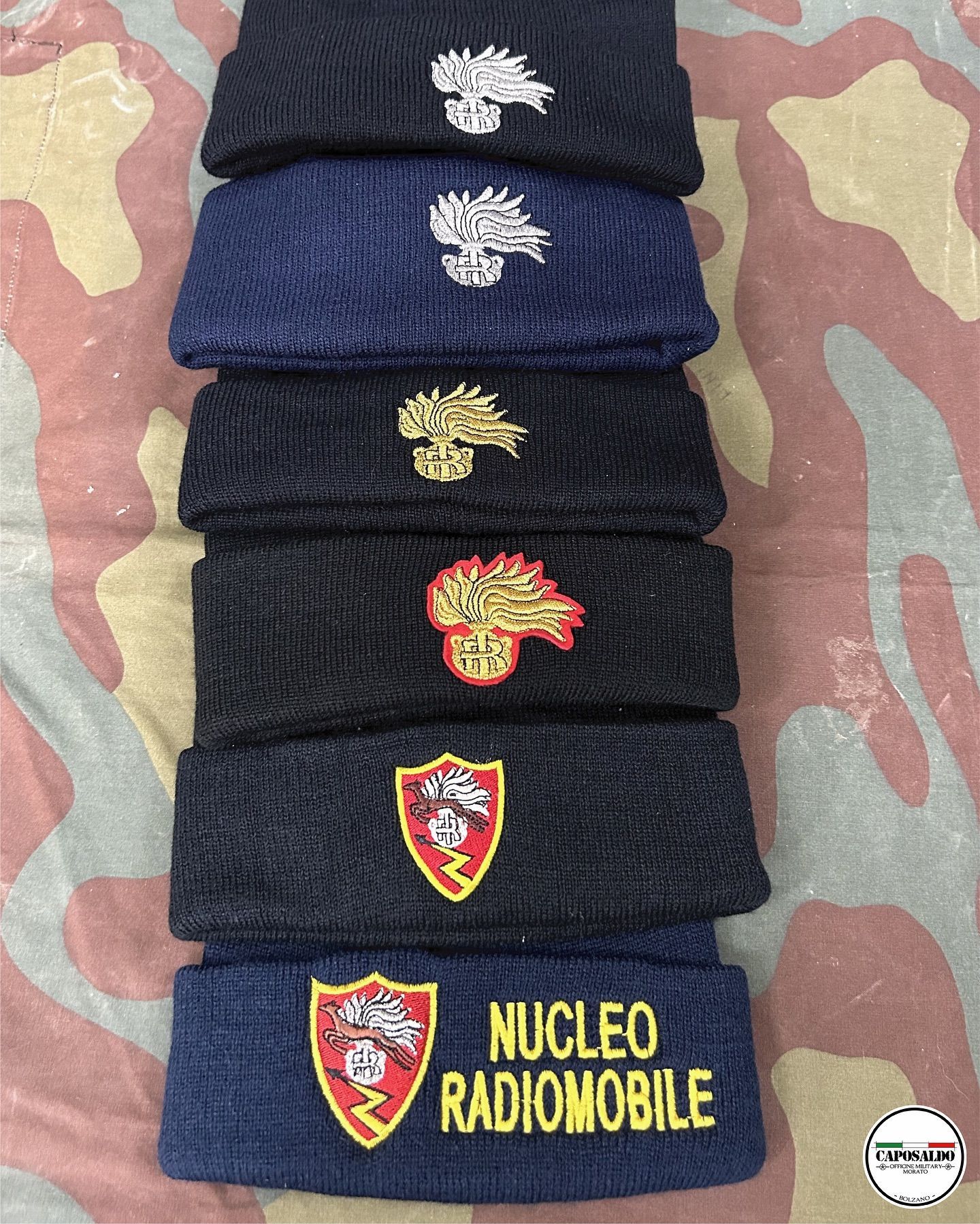 Pila di berretti di lana scura con distintivi ricamati in stile militare, uno dei quali reca la scritta