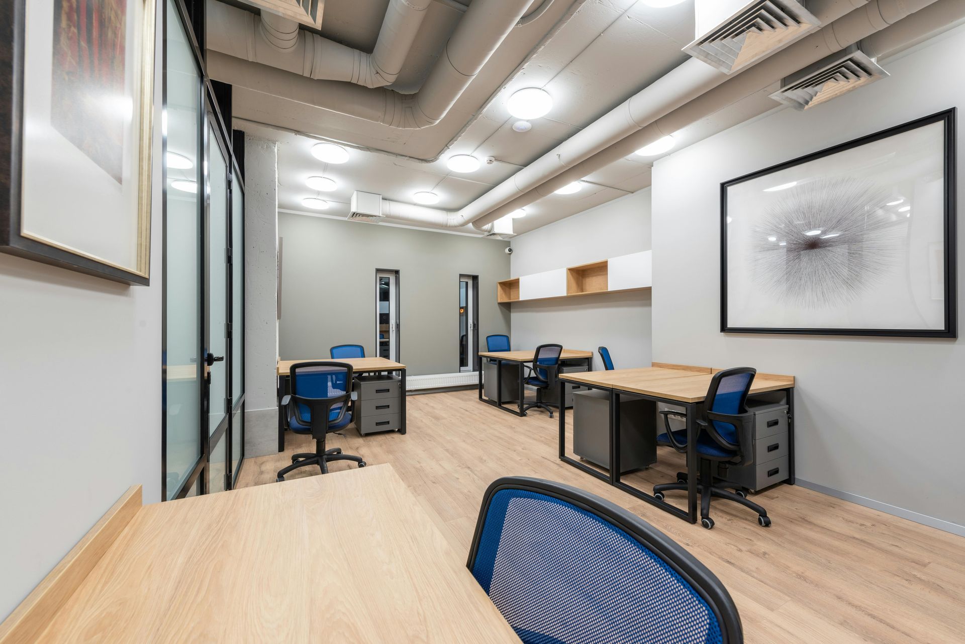 Office space with desks, blue chairs, wood floors, and art on the walls.