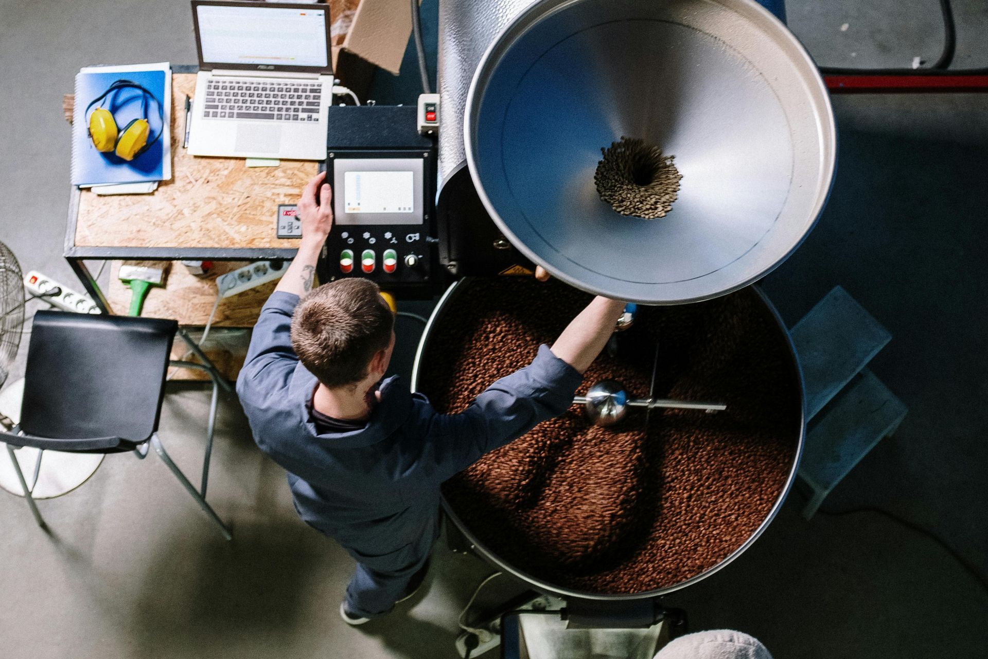 Man roasts coffee beans in a large machine, operating controls. Workshop setting with laptop and tools.