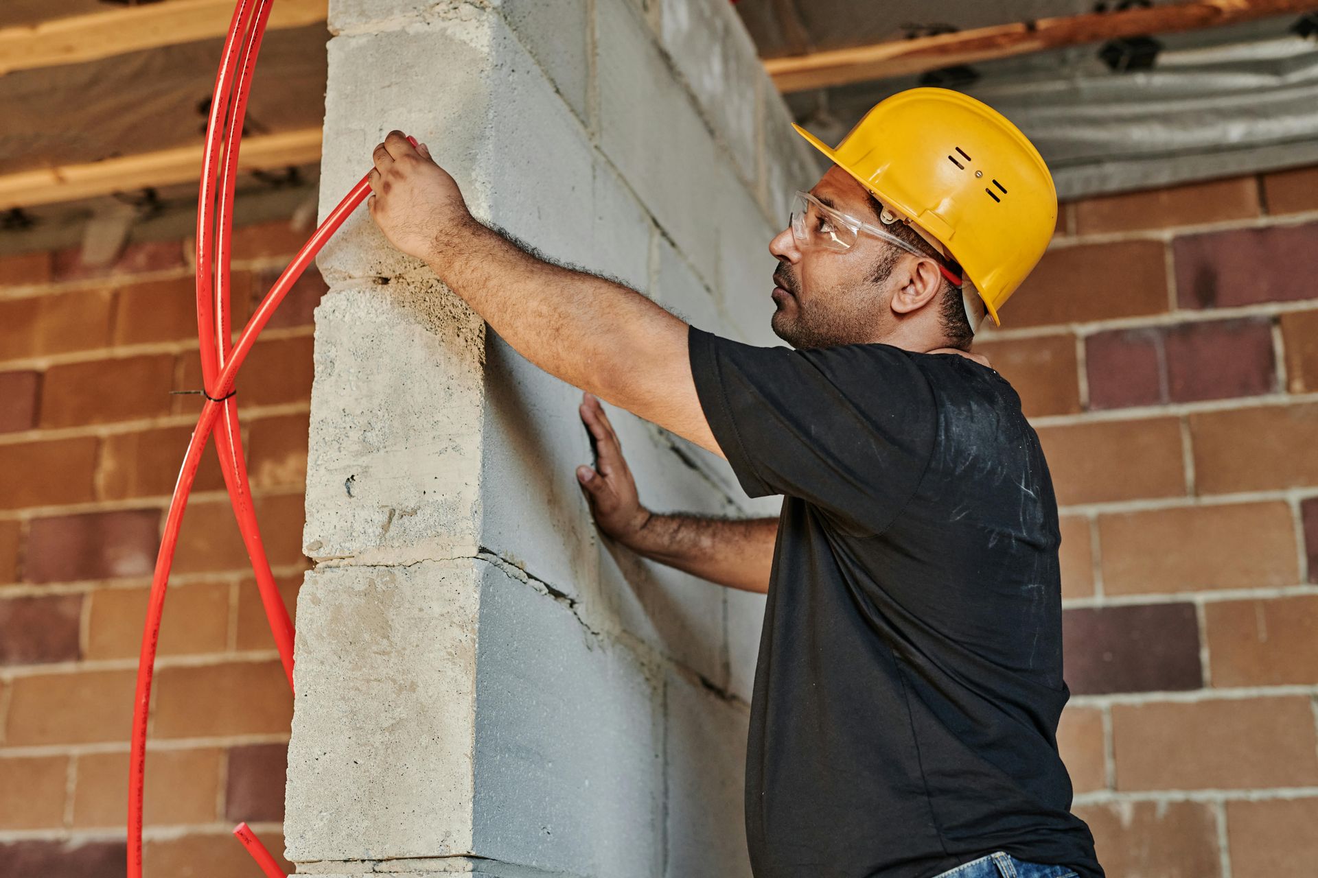 Construction worker in a yellow hard hat and safety glasses, installing red pipes on a cinder block wall.