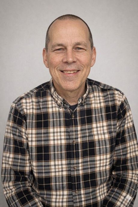 Man with short hair smiling, wearing a plaid shirt, against a gray backdrop.