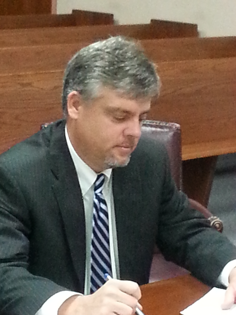 Man in a dark suit writing at a desk in a courtroom or meeting room, head down and focused