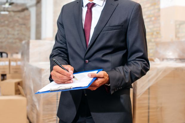 Businessperson in dark suit writing on a clipboard at a warehouse aisle