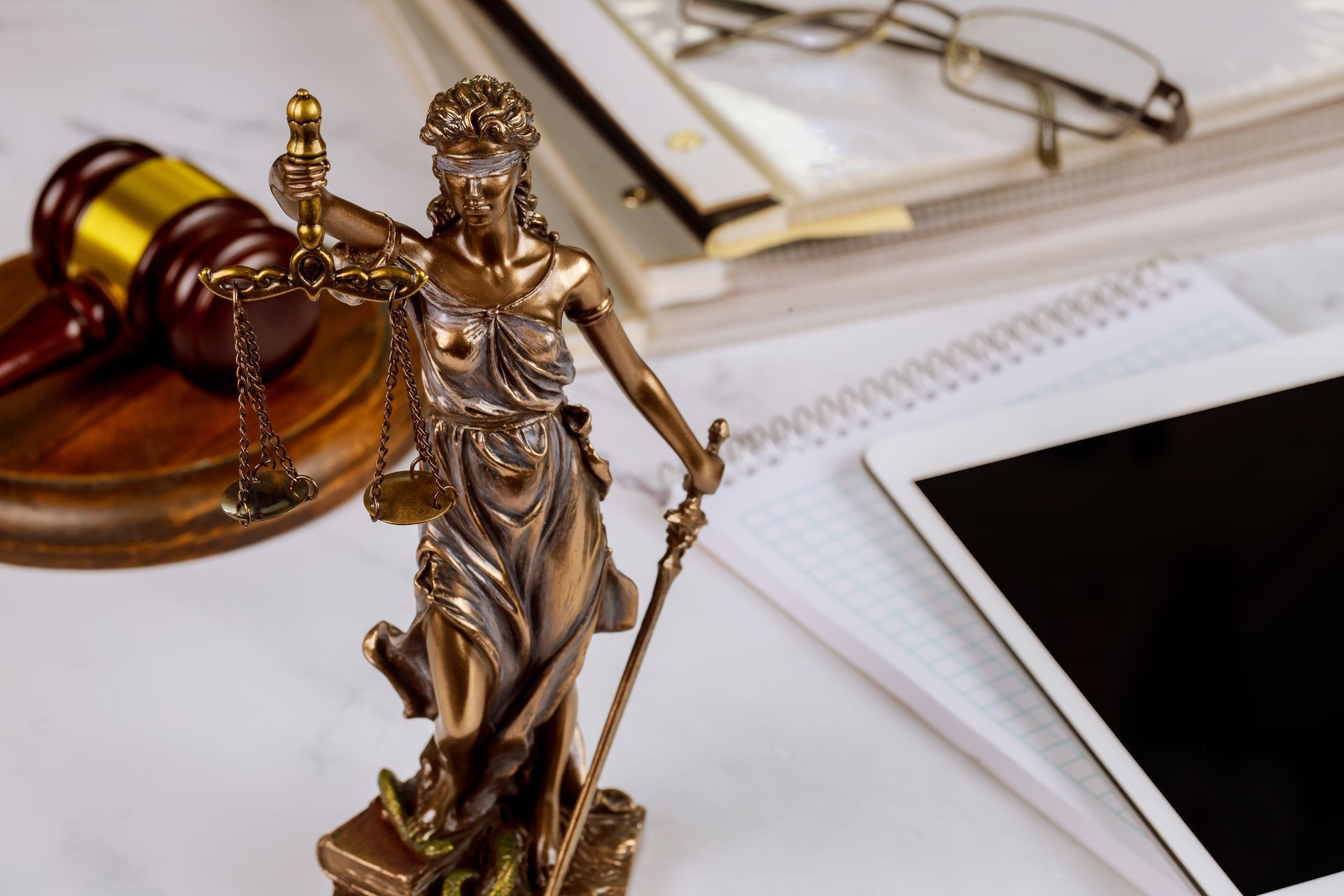 Lady Justice statue beside a gavel, open book, glasses, and tablet on a desk