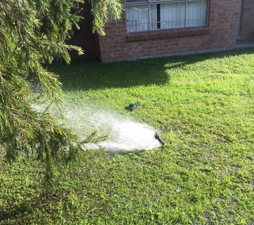 Broken Pipe Spraying Water on a Green Lawn Beside a Brick House — DSR Plumbing Pty Ltd in Ashtonfield, NSW