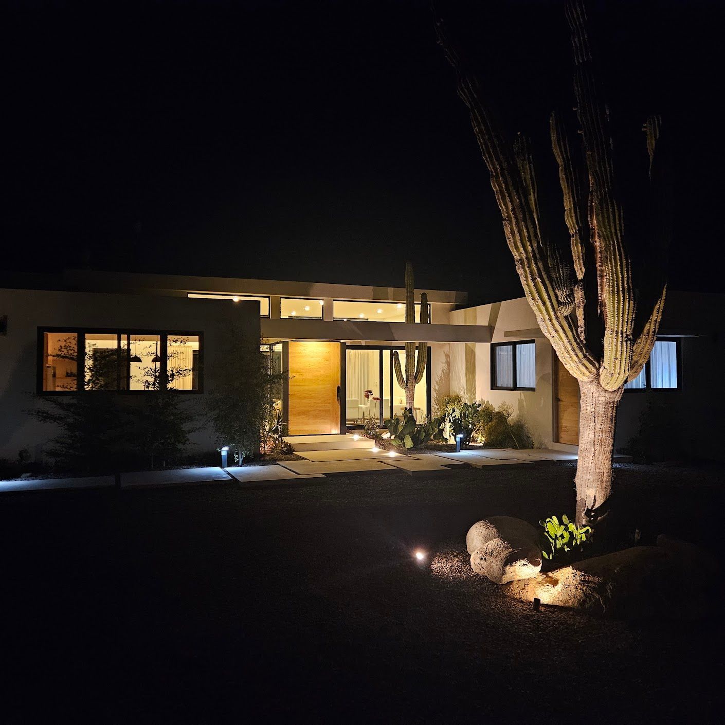 Vista nocturna de una casa moderna iluminada con ventanas resplandecientes y un gran árbol en el patio.