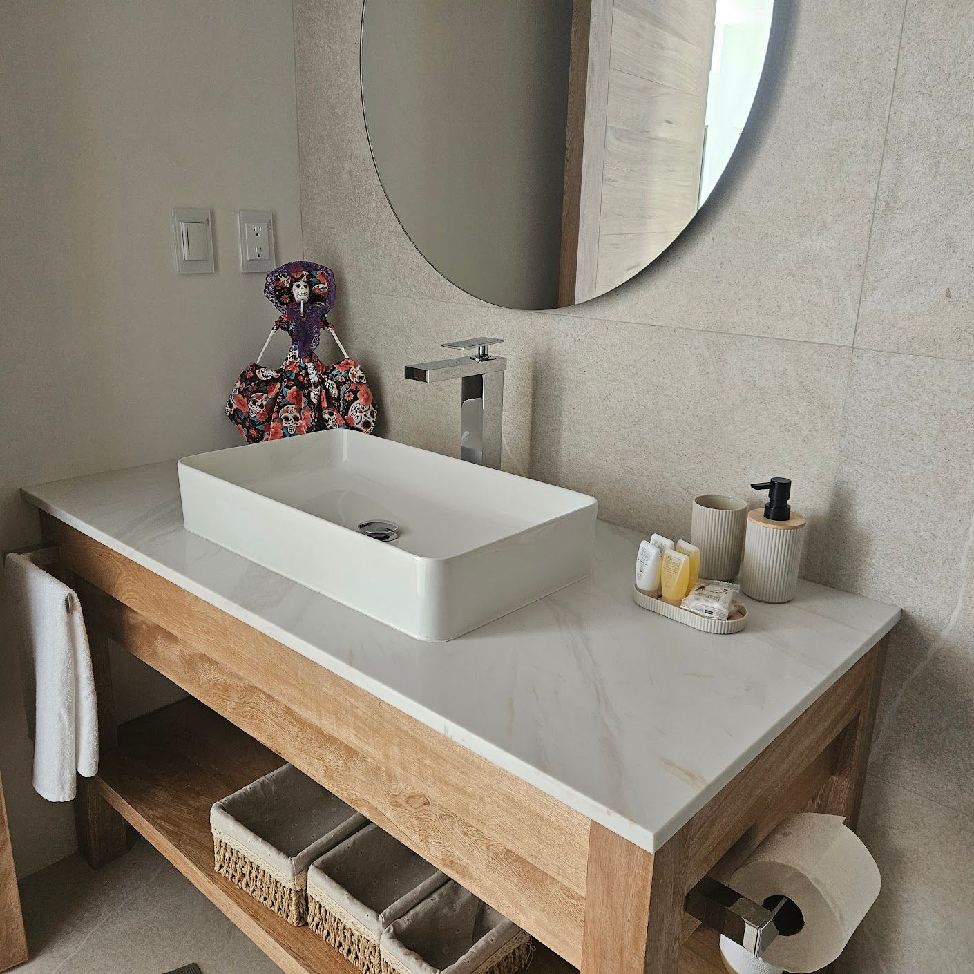 Tocador de baño moderno con lavabo blanco sobre encimera, espejo redondo y toallas sobre un mostrador con marco de madera.
