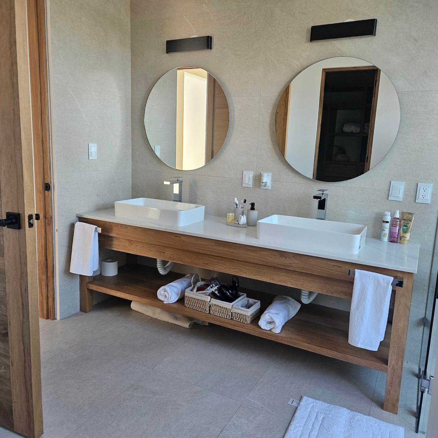 Baño moderno con lavabos dobles, espejos redondos y un tocador de madera con toallas y artículos de aseo.