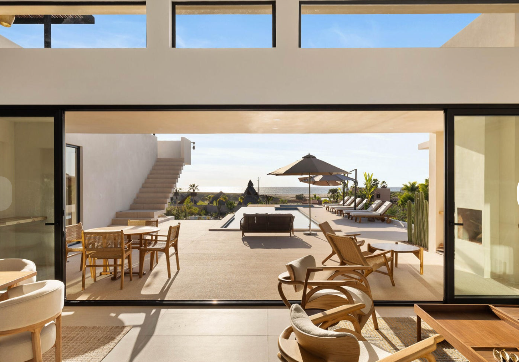 Patio moderno con piscina, tumbonas, mesa de comedor y vista al mar a través de puertas corredizas abiertas.