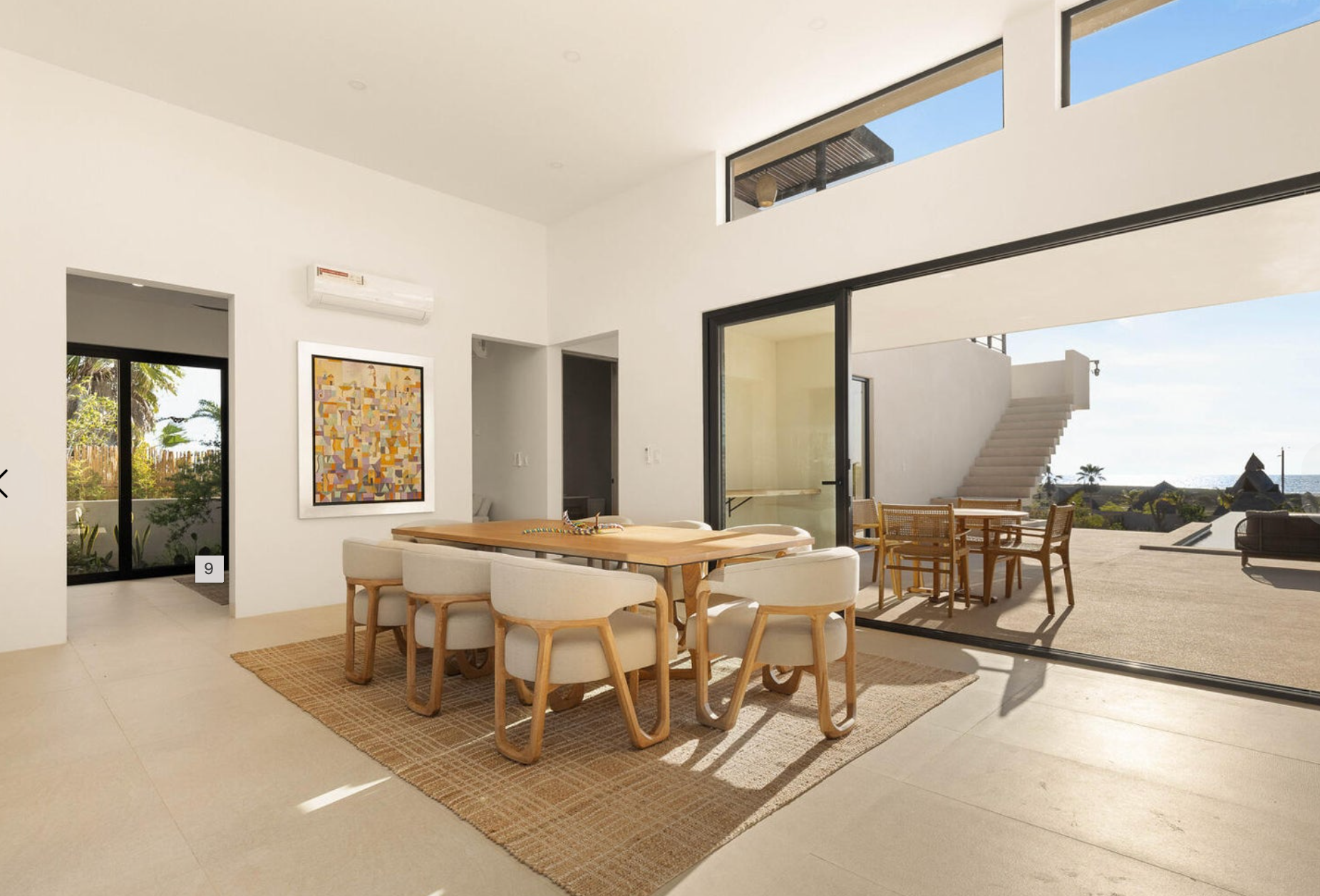 Comedor moderno y luminoso con mesa de madera, sillas de mimbre y puertas correderas que dan a un patio soleado con vistas al mar.