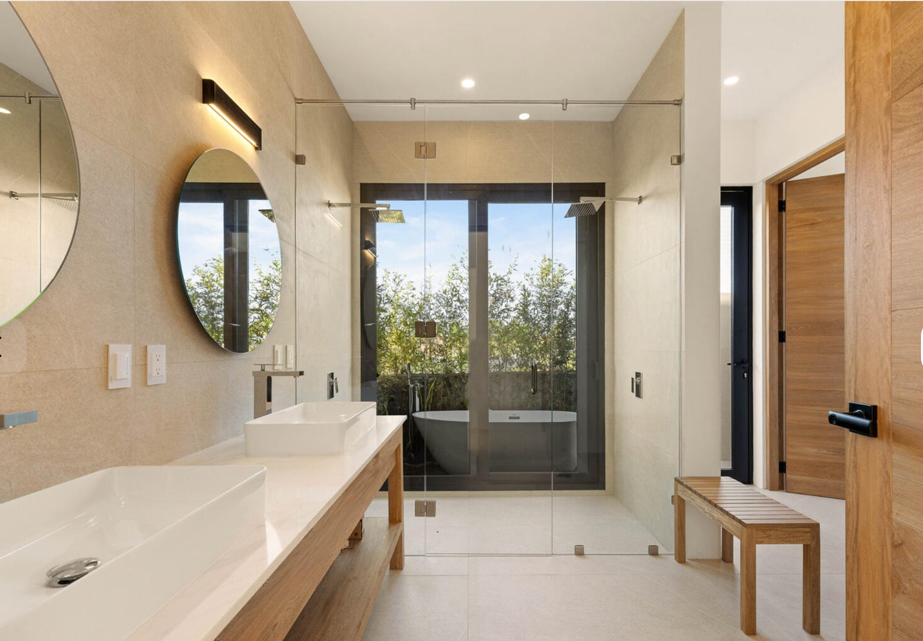 Baño moderno con lavabo doble, espejo redondo, bañera independiente y grandes puertas de ducha de cristal.