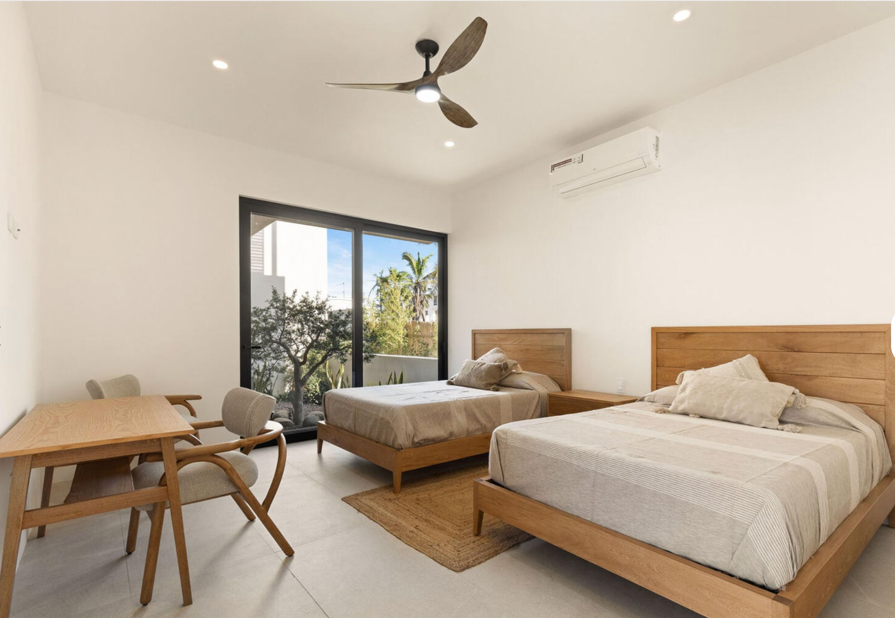 Habitación luminosa con dos camas de madera, mesa y sillas, ventilador de techo y puertas que dan al balcón.