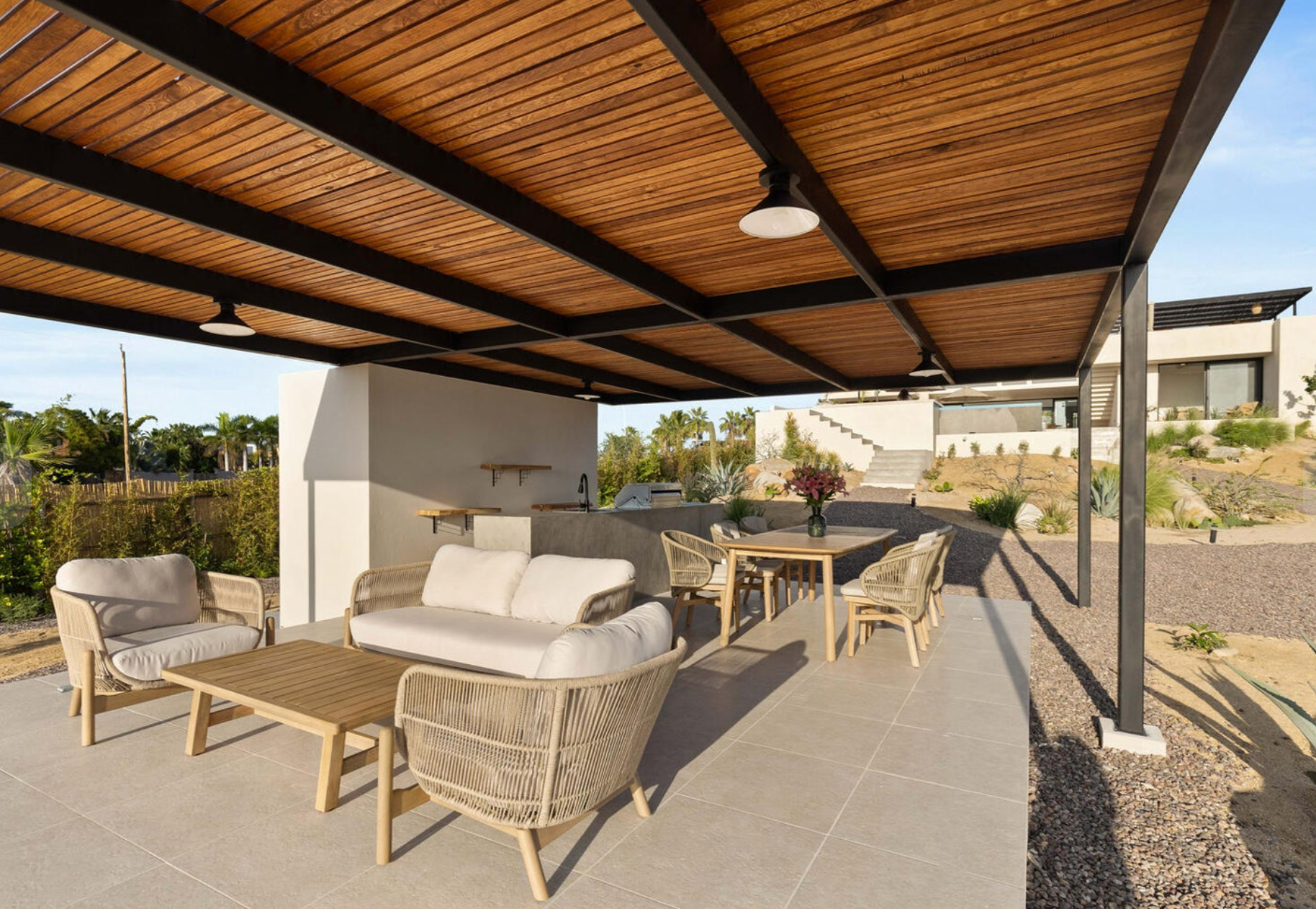 Zona de estar cubierta en el patio con sillas de mimbre y muebles de salón bajo un techo con vigas de madera.