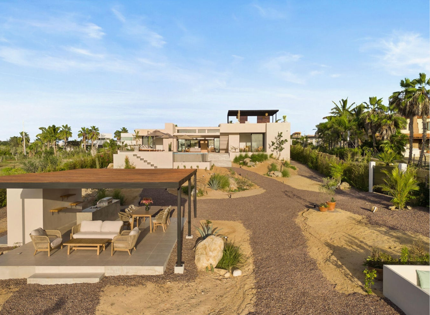 Patio de un complejo turístico en el desierto con asientos a la sombra, sendero serpenteante, palmeras y villas modernas bajo un cielo azul.