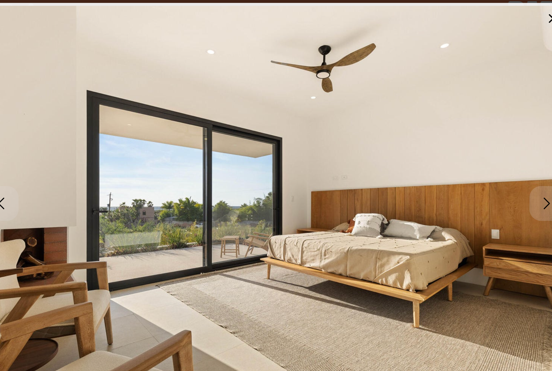 Dormitorio moderno con una gran puerta corrediza de vidrio, cama, silla de madera y ventilador de techo con vistas a la vegetación.