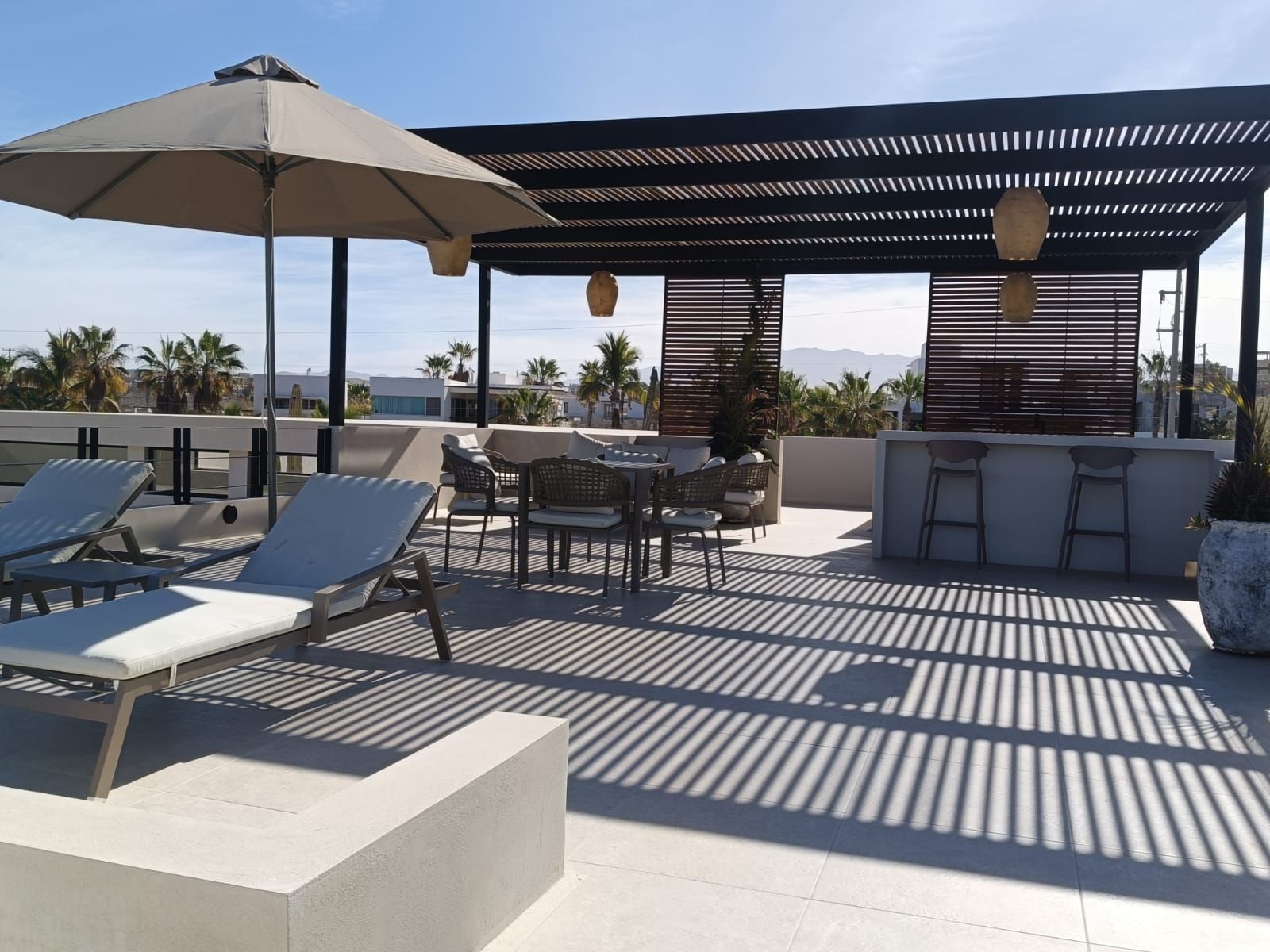 Terraza en la azotea con tumbonas, sombrilla y barra a la sombra bajo una pérgola, disfrutando del sol.