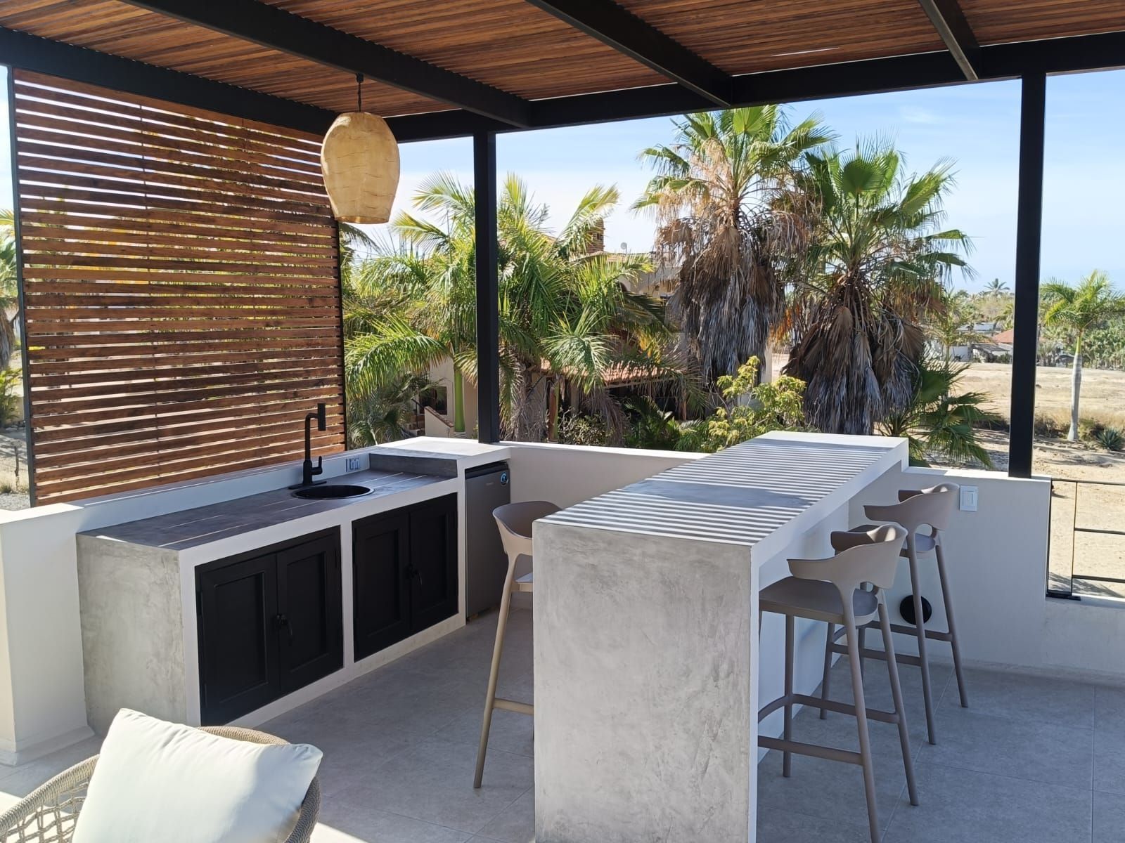 Cocina exterior en el patio con isla de hormigón, taburetes y palmeras más allá de una mampara de bambú.