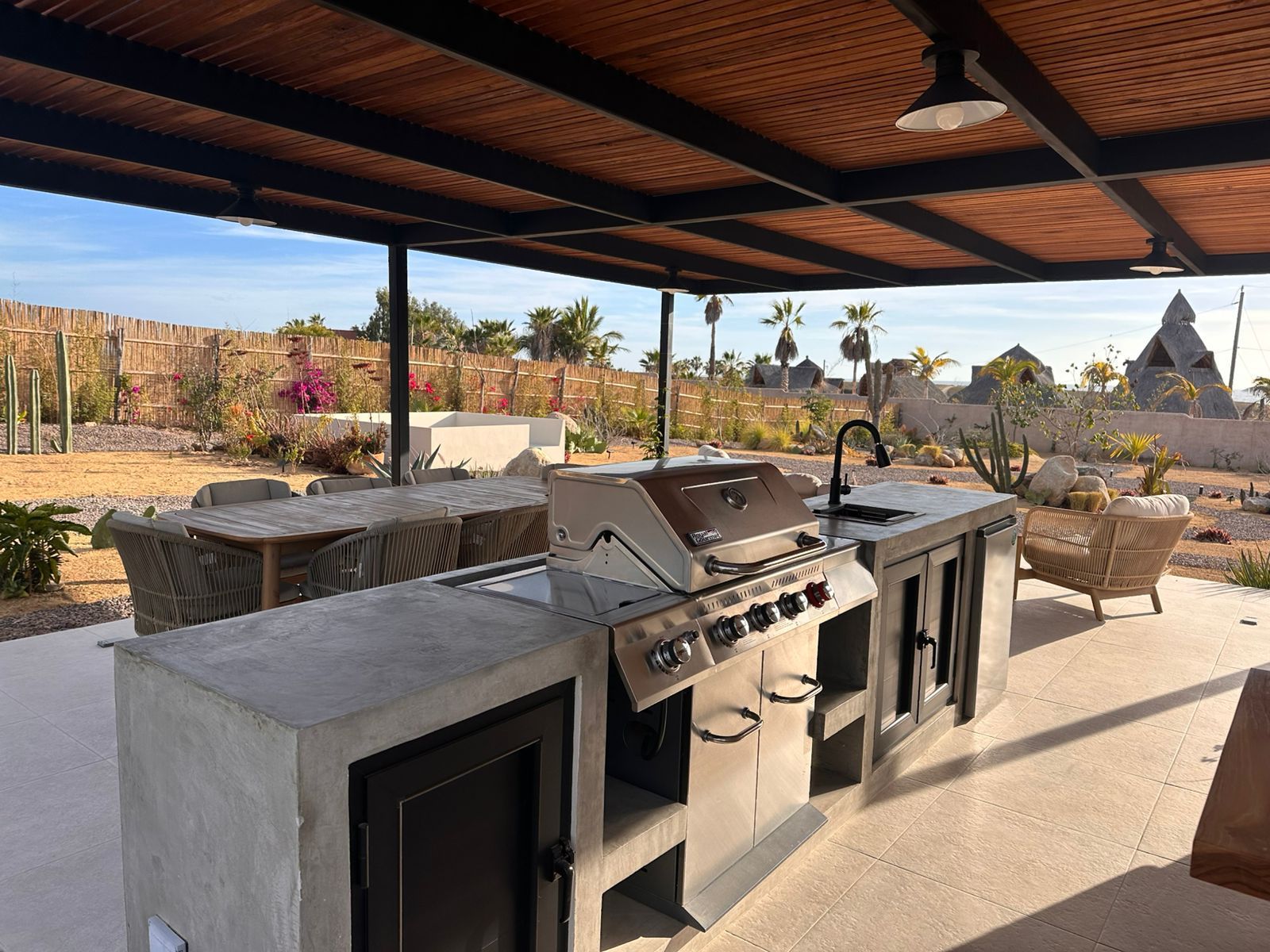 Cocina exterior con patio, parrilla, fregadero y encimera bajo un techo de madera, con vistas a un paisaje desértico.