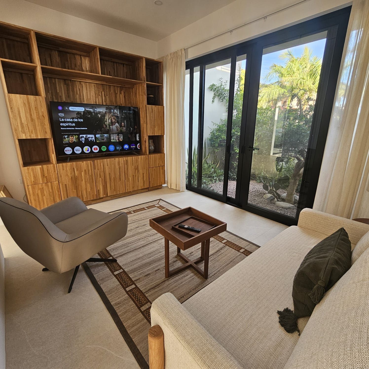 Sala de estar moderna con pared para TV, sillón, sofá, mesa de centro y grandes puertas de cristal que dan a un jardín.