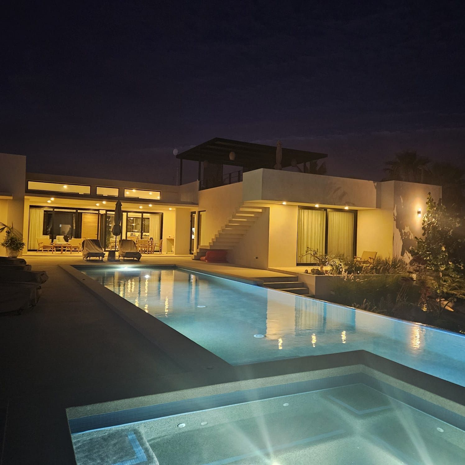 Villa moderna de noche con piscina iluminada y jacuzzi que refleja luces cálidas.