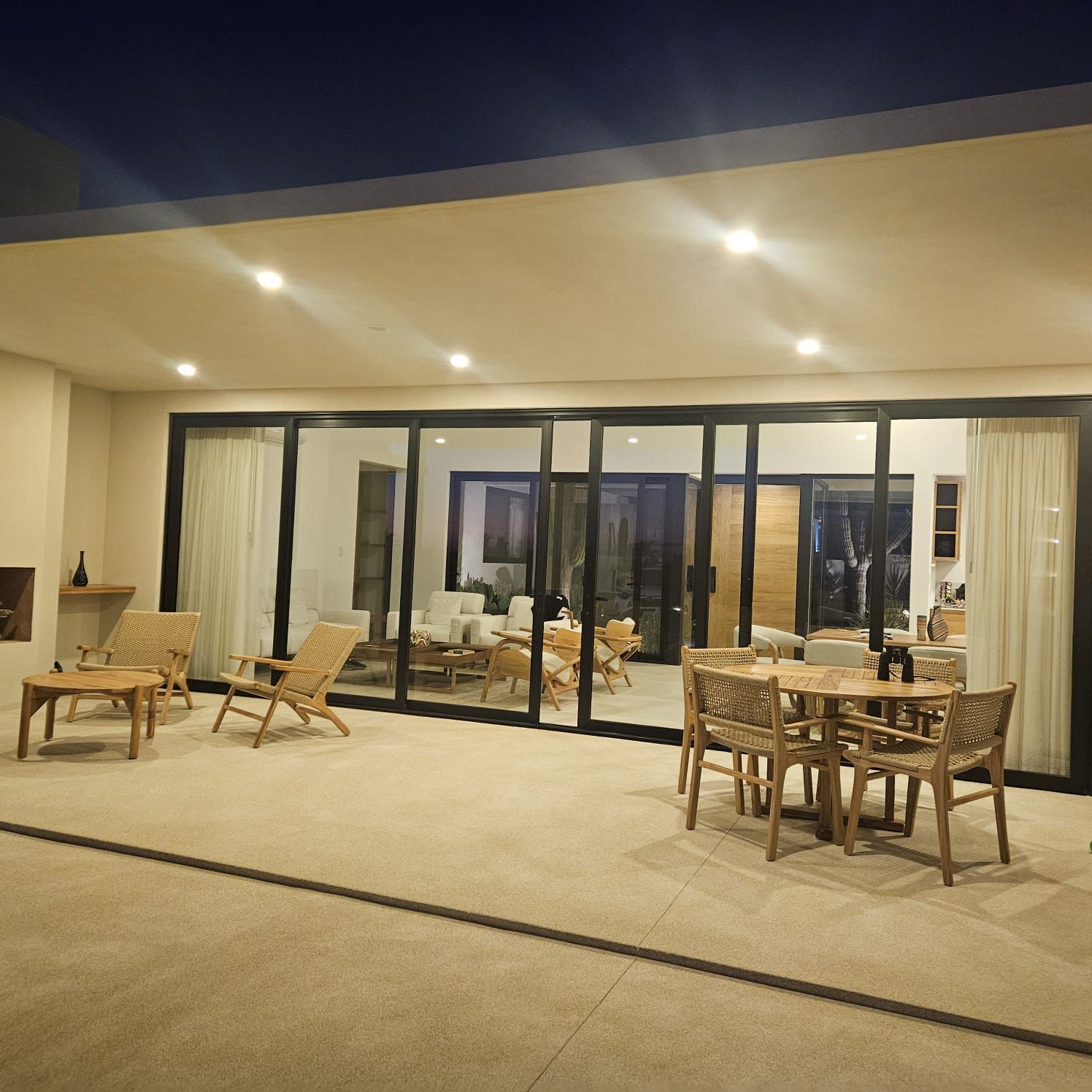 Patio exterior con mesas y sillas de mimbre junto a un edificio con paredes de cristal por la noche.