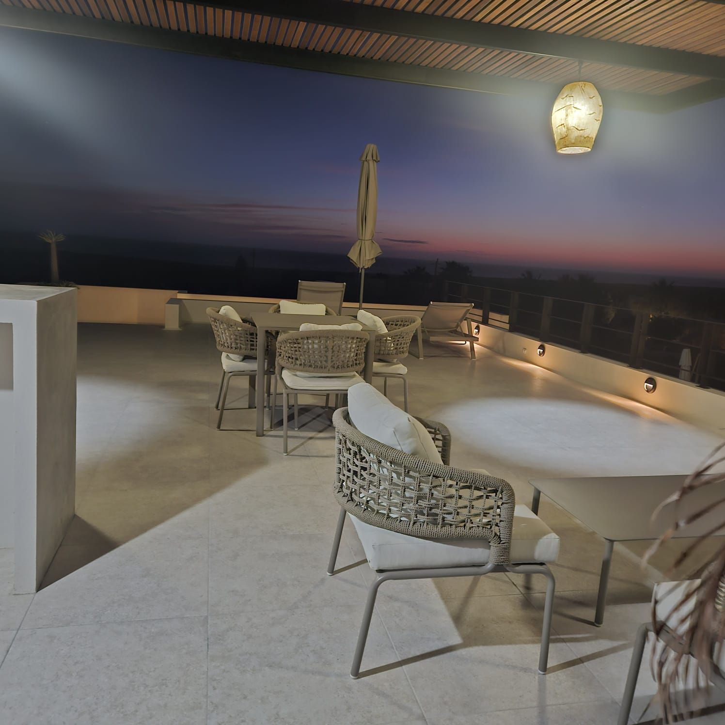 Terraza al aire libre con mesas y sillas al atardecer, con vistas a un horizonte tranquilo bajo un cielo de suaves tonos azules y rosados.