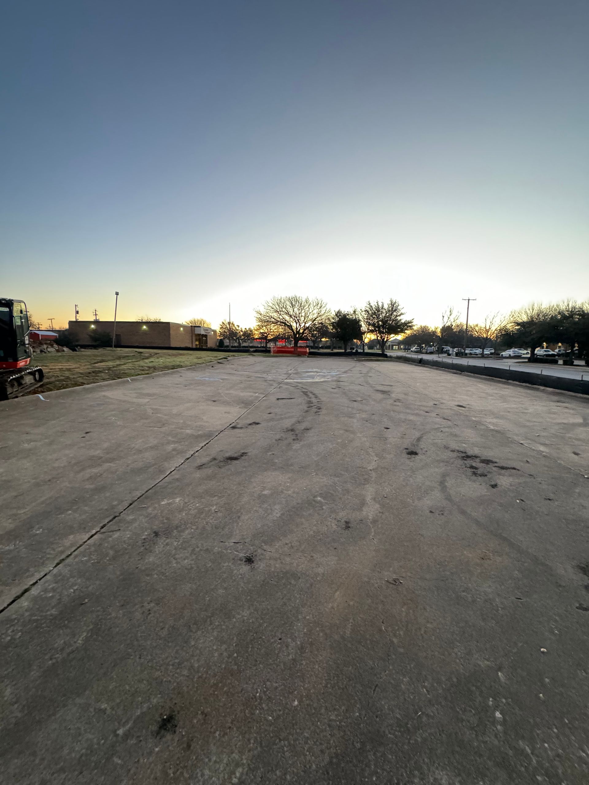 An empty, cracked asphalt lot at dawn with trees and buildings in the background.