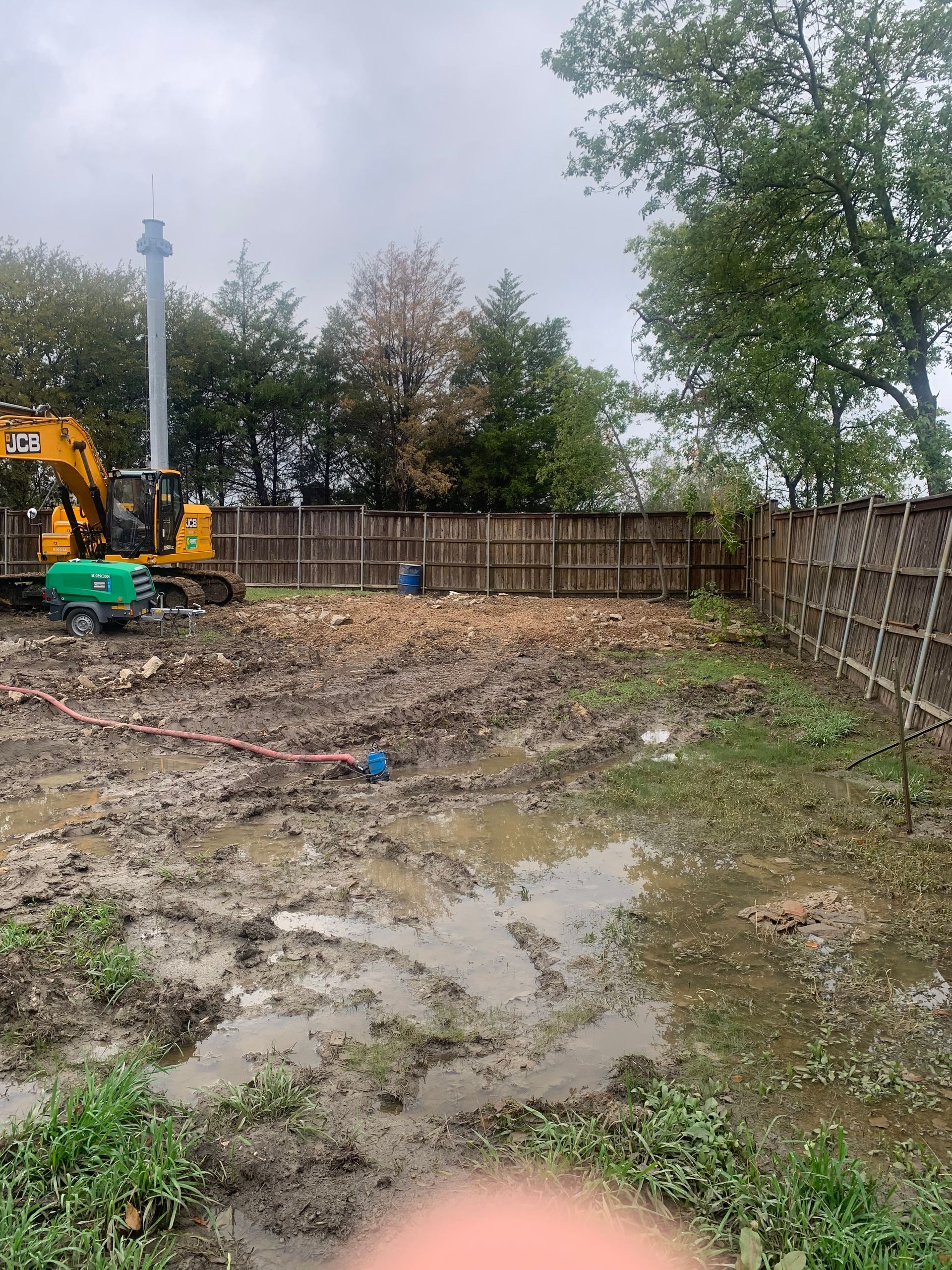 Muddy construction site with excavator, wooden fence, and puddles under overcast sky.