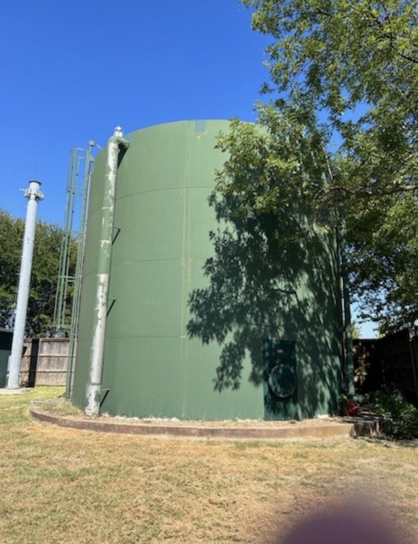Green cylindrical water tank outdoors next to a ladder and trees under a blue sky.