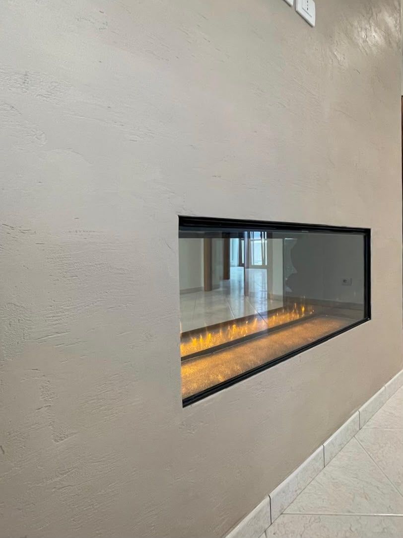 Stretta finestra di vetro incassata in una parete beige, da cui si intravede un ambiente interno illuminato.