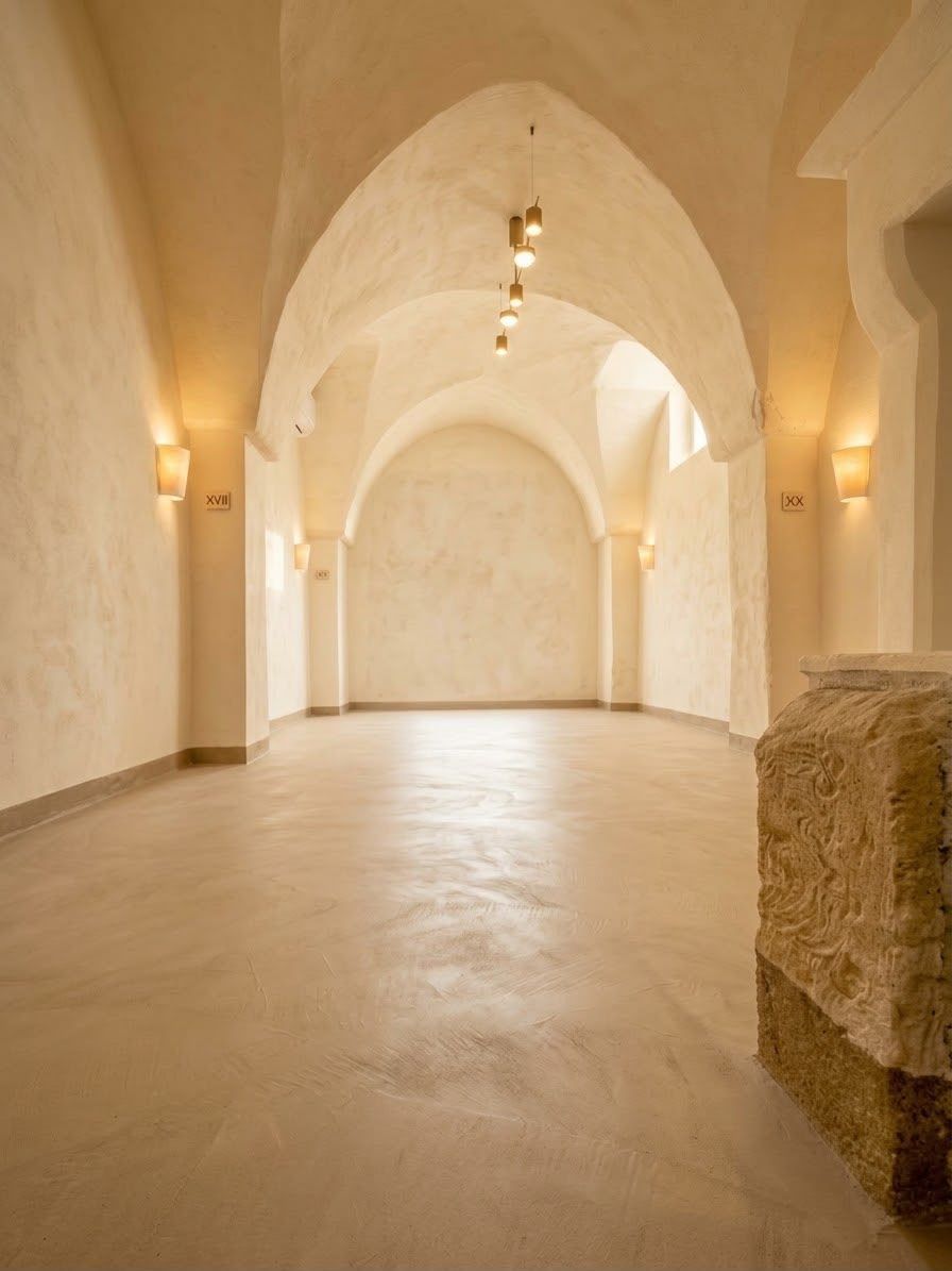 Corridoio ad arco color beige caldo, con pareti in pietra e luci soffuse alle pareti, visto dall’interno di un atrio vuoto