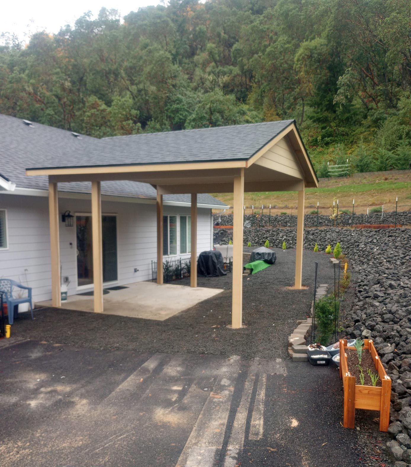 Wooden carport beside a house on a paved driveway, with a rocky retaining wall and wooded hillside behind.