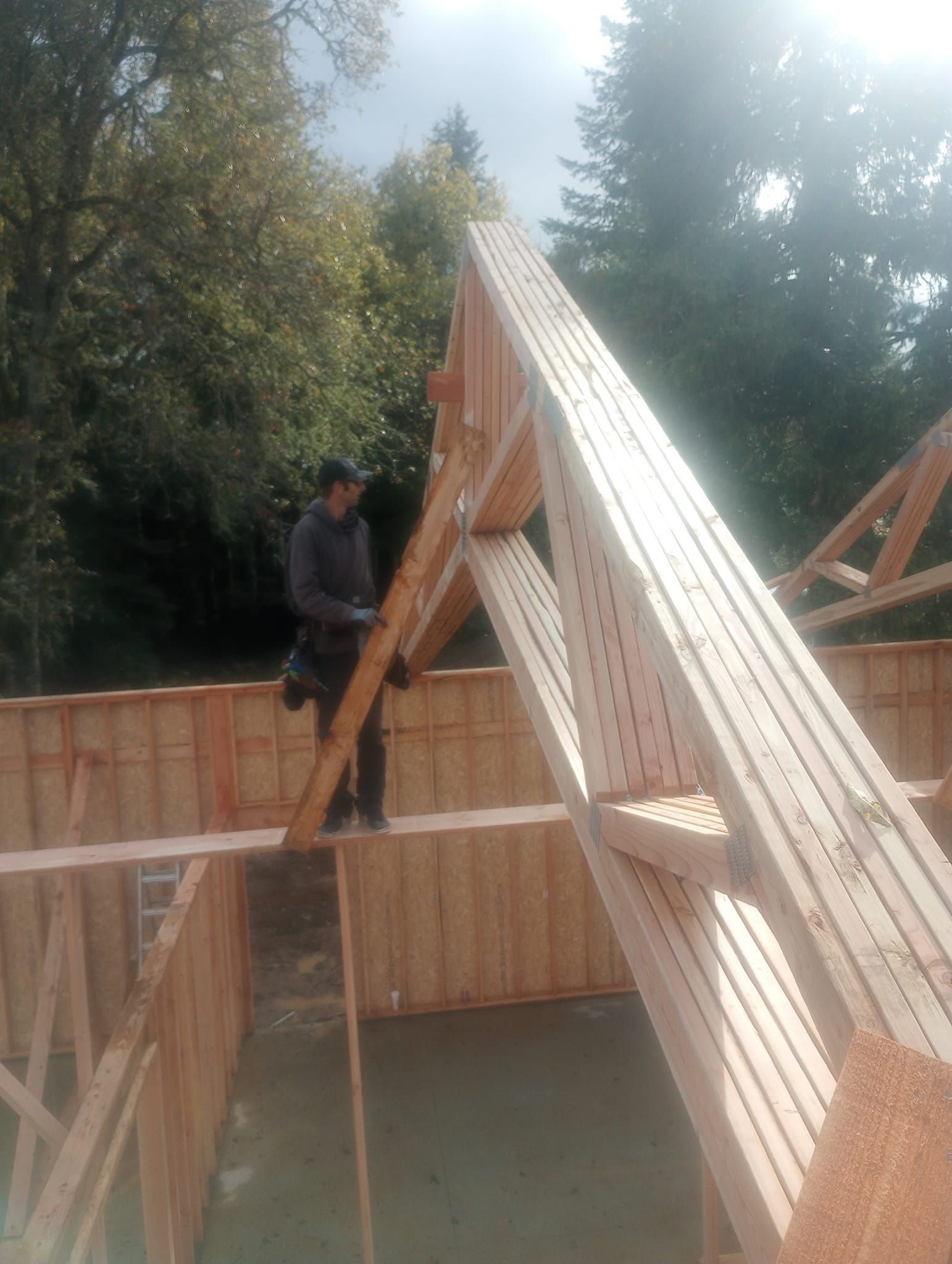Worker on a wooden roof truss at a construction site, with timber walls and trees in the background.