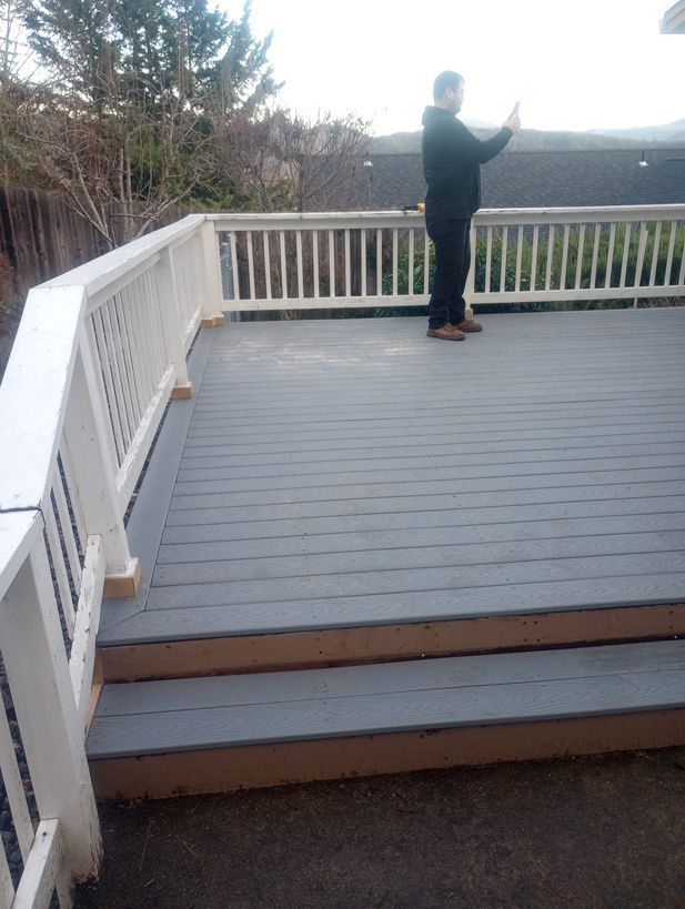 Person standing on a gray wooden deck, holding up a phone near white railings outdoors.