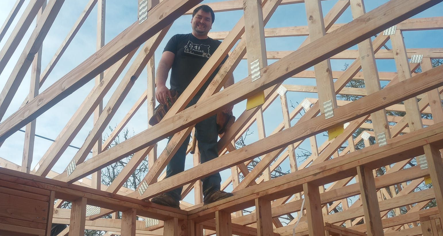 Person standing on a wooden roof frame at a construction site with blue sky behind.