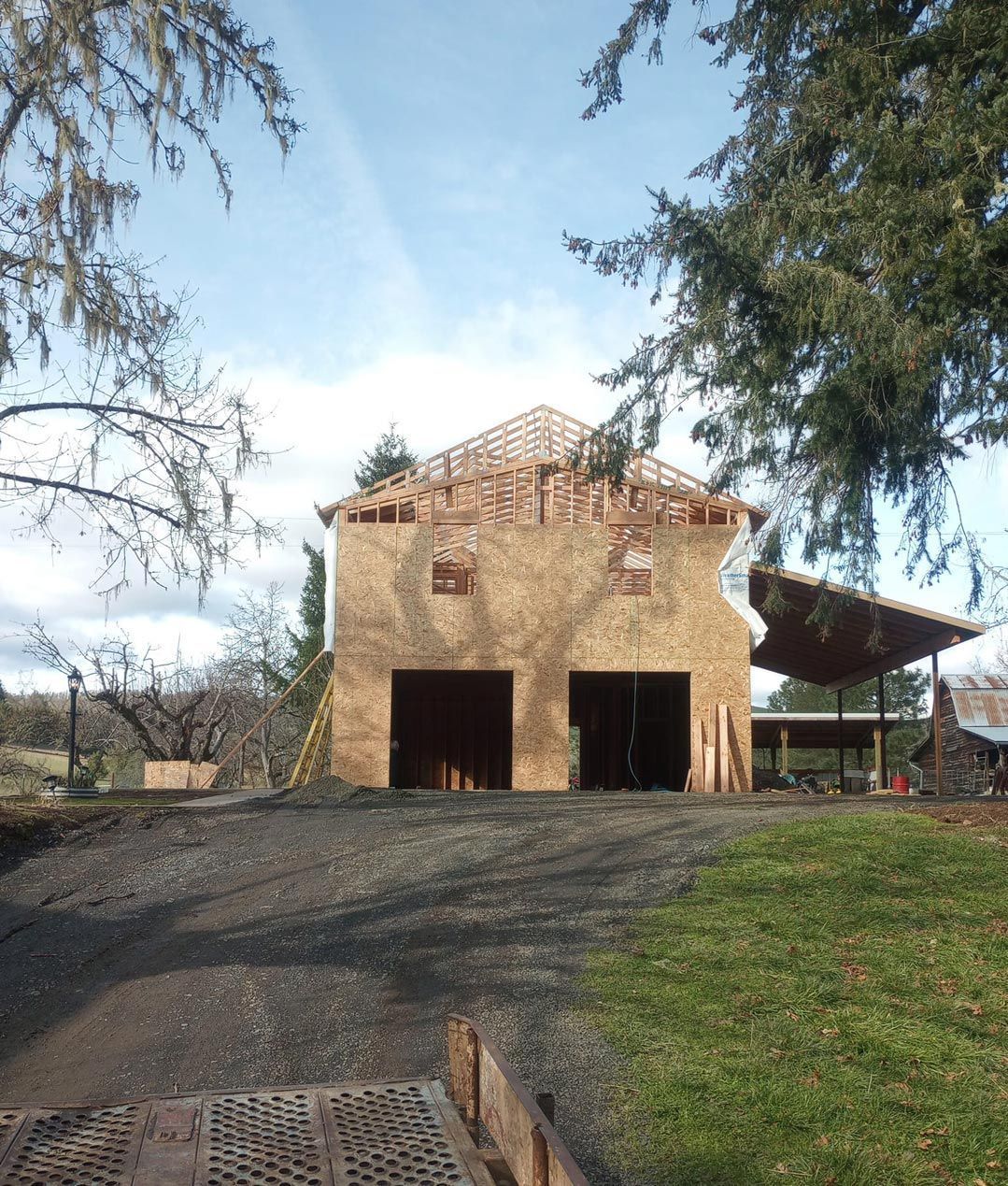 Two-story tan barn with open garage bays on a gravel driveway, framed by trees under a blue sky.