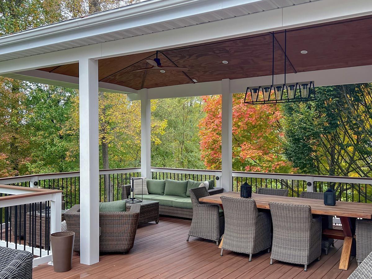 Covered outdoor patio with wicker seating, dining table, and autumn trees beyond the railing