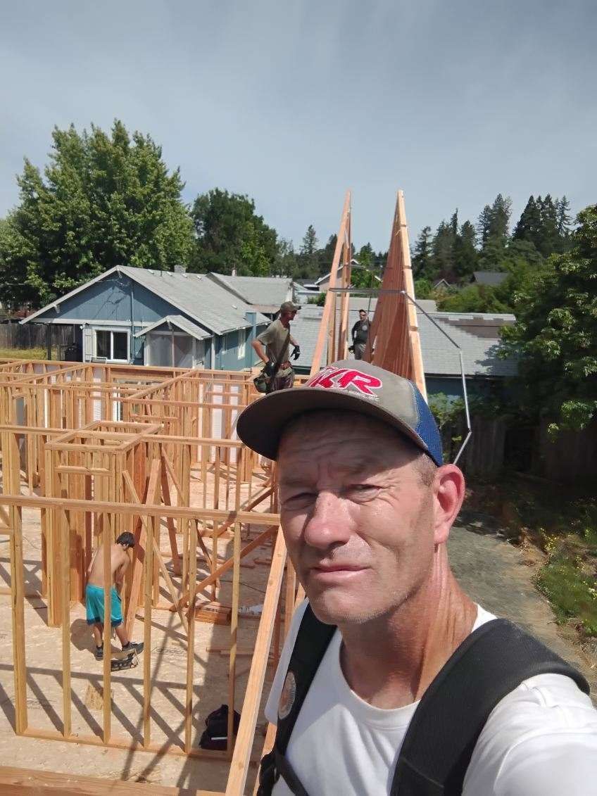 Man in black cap taking selfie at a house construction site with wooden framing and workers behind him