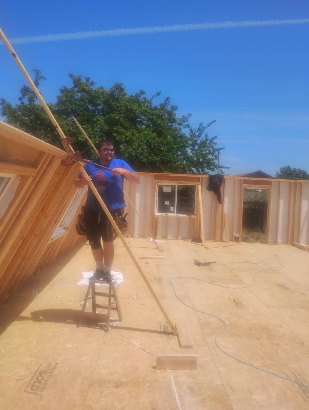Worker on a stool stretching a beam at a wooden house construction site under a blue sky