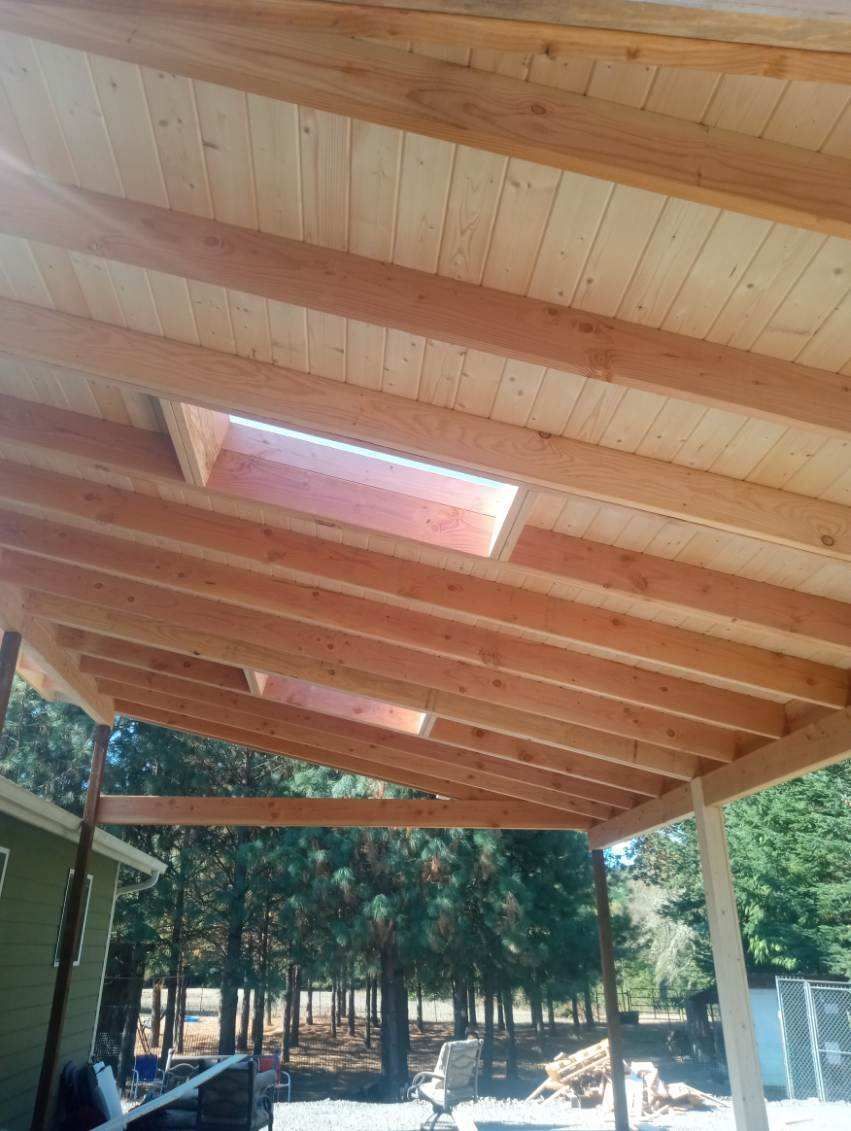 Wooden pavilion roof with exposed beams and skylights over an outdoor forested area.