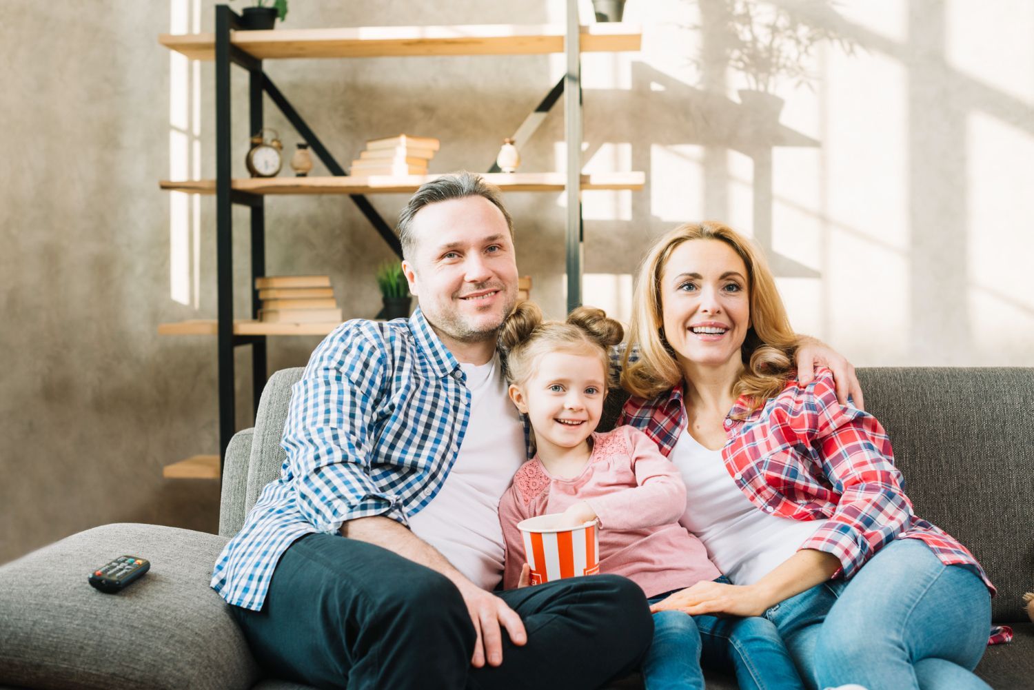 Family sitting on a couch, smiling in a bright living room with shelves behind them.