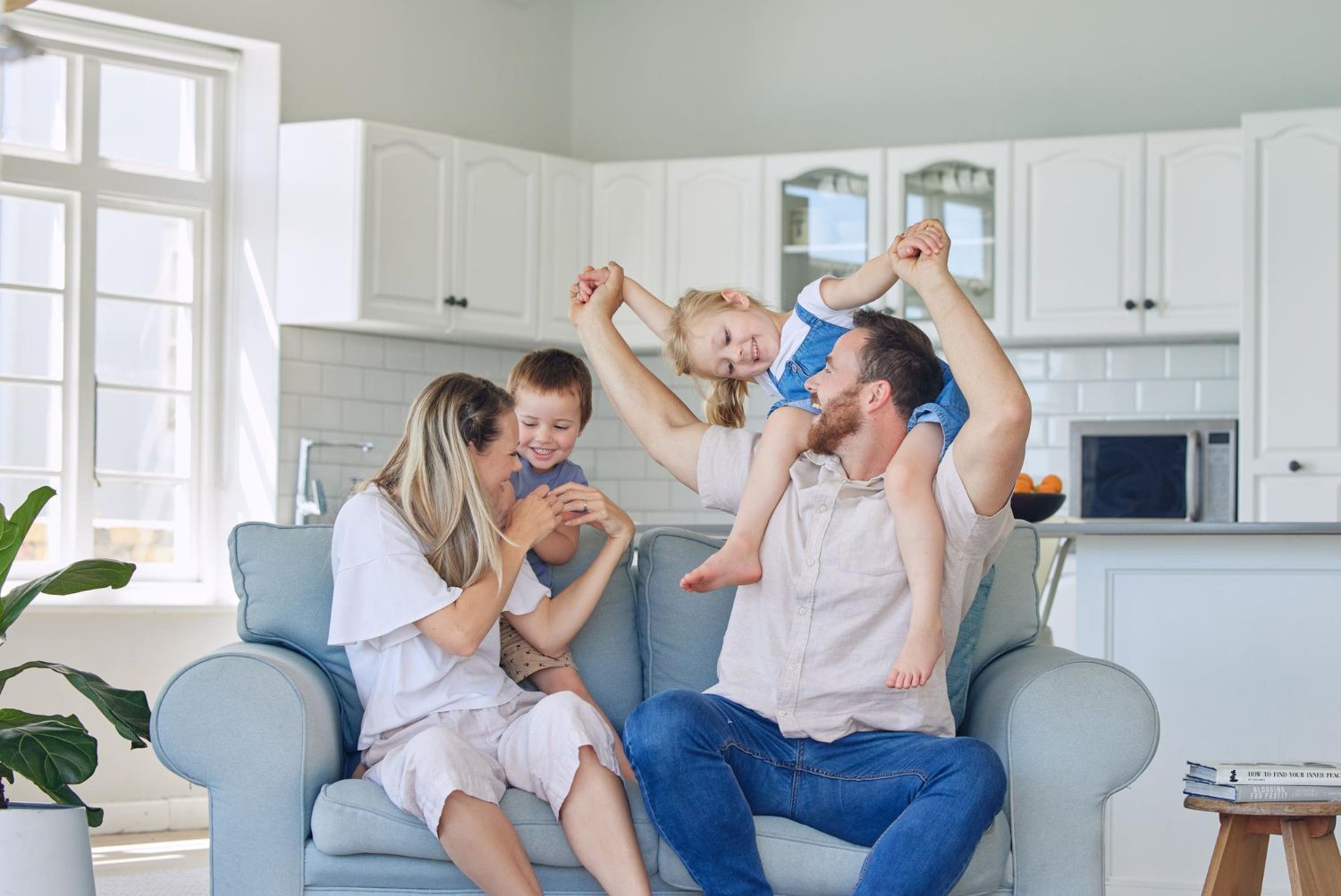 Family sitting on a blue couch in a bright kitchen, laughing and playing with two young children